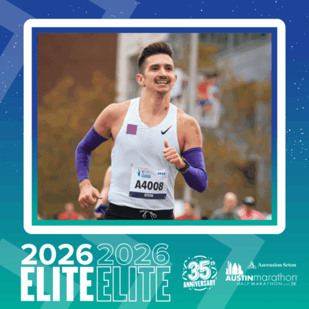A male runner in a white tank top and purple arm sleeves smiles as he crosses the finish line. His bib reads 