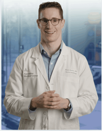 A smiling doctor wearing glasses and a white lab coat stands with hands clasped, ready for registration. The coat reads "Ascension Medical Group" and "Ryan Quinn, MD, Sports Cardiology." The background appears to be a medical or hospital setting. Austin Marathon Half Marathon & 5K
