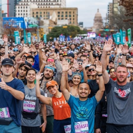 A large group of marathon runners stand packed together at the starting line, many smiling and raising their hands enthusiastically, embodying the energy needed for future trends and strategic planning. Buildings and trees line the street in the background. Austin Marathon Half Marathon & 5K
