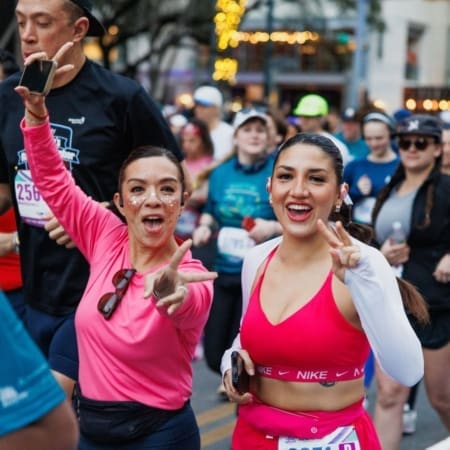 Two women in athletic wear smile and gesture enthusiastically at the camera during a crowded outdoor race event. Other runners fill the background, and festive lights glow in the trees—a glimpse of future trends in fitness for what's next in 2027. Austin Marathon Half Marathon & 5K