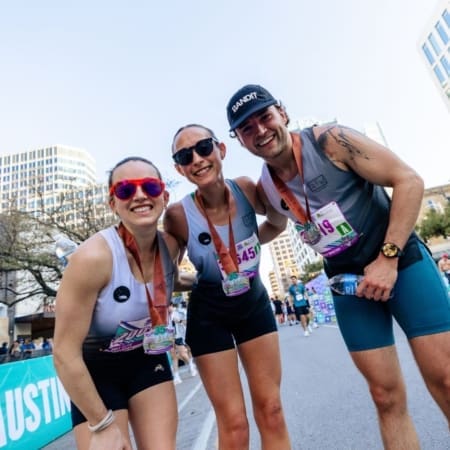 Three smiling runners wearing medals and race bibs pose together on a city street after finishing a race. Tall buildings and other participants hint at the exciting future trends for city marathons leading up to 2027. Austin Marathon Half Marathon & 5K