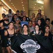 A group of smiling runners from social teams pose together indoors, holding a black "SEXPYPACE" banner with a disco ball graphic. Most are in athletic wear, some make peace signs, and the cheerful atmosphere reflects their shared training spirit. Austin Marathon Half Marathon & 5K