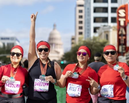 Four women from non profit teams in matching red outfits and medals smile after a race, proudly holding up their race bibs. One woman raises her arm victoriously with the Texas State Capitol visible in the background. Austin Marathon Half Marathon & 5K