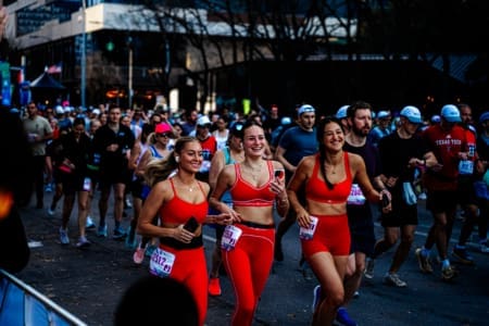 A group of runners participate in a race, with three women in matching bright red outfits—likely part of social teams—smiling and running together in the foreground. Other runners fill the street behind them, showing their dedication to training. Austin Marathon Half Marathon & 5K