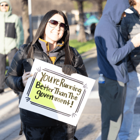A smiling person wearing sunglasses and a black jacket holds a sign that reads, "You’re running better than the government!" at an outdoor event, surrounded by enthusiastic spectators and Austin’s crowd in the background. Austin Marathon Half Marathon & 5K