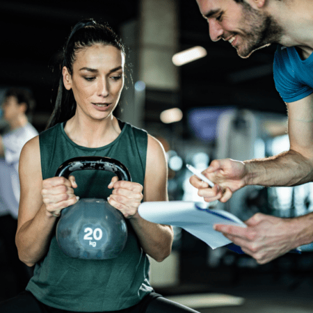 A woman holding a 20 kg kettlebell sits while a man beside her smiles and shows her a clipboard with papers, likely discussing her Strength Training for Runners plan in preparation for the Austin Marathon in a gym setting. Austin Marathon Half Marathon & 5K