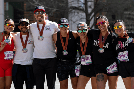 Six smiling runners stand together outdoors, wearing sunglasses, athletic gear, race bibs, and finisher medals around their necks, celebrating as charity teams after completing a race. Austin Marathon Half Marathon & 5K