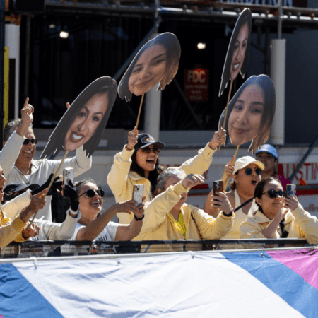 A group of spectators in light-colored clothing cheer and hold large cutouts of a woman's face while taking photos at an outdoor event, creating an electric atmosphere against the backdrop of a construction site. Austin Marathon Half Marathon & 5K
