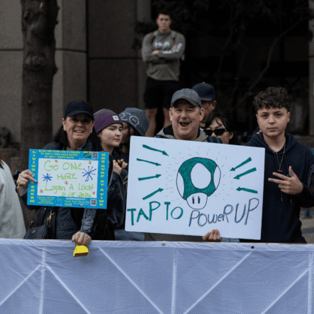 A group of spectators stand behind a barrier at an outdoor event, smiling and holding signs. One sign reads "Go ONE MORE Logan & West" and the other features a Mario-style mushroom and says "TAP TO Power UP. Austin Marathon Half Marathon & 5K