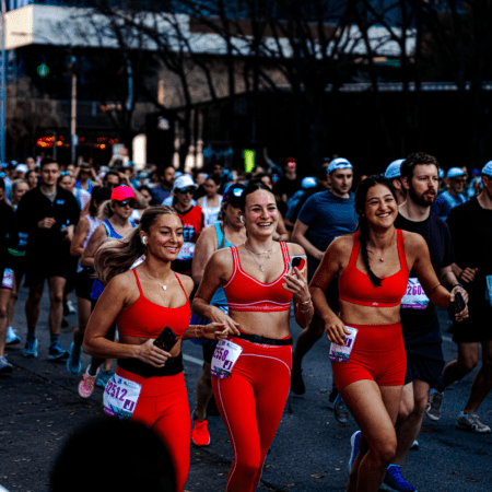 A group of marathon runners is on a city street. Three women in matching red athletic outfits smile and run together in the foreground, surrounded by an enthusiastic Austin crowd. It's a lively, energetic scene full of Austin spirit. Austin Marathon Half Marathon & 5K