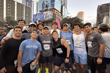 A group of twelve young men from collegiate teams wearing race bibs and athletic clothes smile together at a city street outdoor running event, with tall buildings and trees in the background. Austin Marathon Half Marathon & 5K