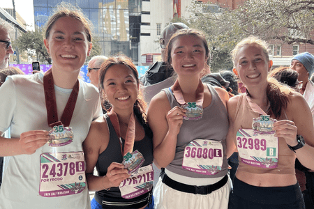 Four smiling women from collegiate teams wearing race bibs and medals pose together after finishing a marathon on a city street. They proudly hold up their medals, dressed in athletic gear, with buildings and trees in the background. Austin Marathon Half Marathon & 5K