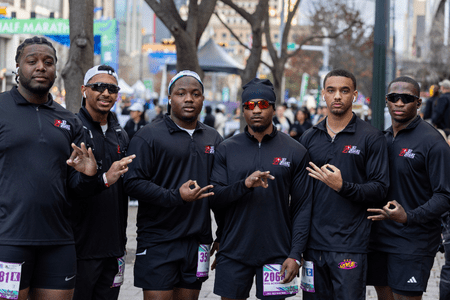 Six men in matching black athletic wear pose together outdoors at a marathon event, each making hand signs, showing off their race bibs—a perfect example of social teams coming together through training, with city buildings in the background. Austin Marathon Half Marathon & 5K