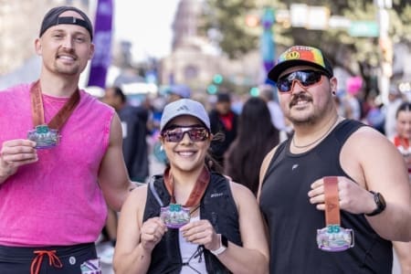 Three runners smile and hold up medals after a race, representing their community teams. They stand outdoors with a crowd and a domed building in the blurred background, wearing athletic gear, sunglasses, and hats. Austin Marathon Half Marathon & 5K