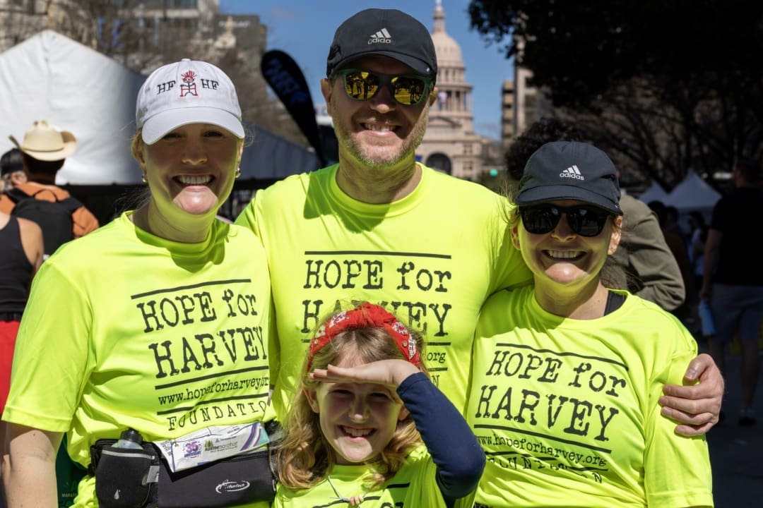 Four people wearing bright yellow "Hope for Harvey" shirts pose and smile outdoors at a Charity Teams fundraising event. A child stands in front, making a hand gesture, with the Texas Capitol visible in the background. Austin Marathon Half Marathon & 5K