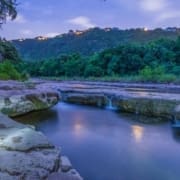 A serene river flows over flat rocks at dusk, reflecting a soft blue sky. Lush green trees line the banks—one of the top 5 running trails among Austin running routes—while houses with glowing lights dot the distant hilltop under a cloudy sky. Austin Marathon Half Marathon & 5K