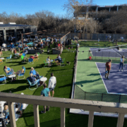 People relax on outdoor chairs and socialize next to a pickleball court, enjoying the sunshine and spring in Austin. Groups mingle on what’s known as one of the best patios for runners, with trees and a building providing a scenic backdrop. Austin Marathon Half Marathon & 5K