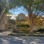 A two-story brick house with a metal roof and large windows is partly shaded by tall trees. Nestled in quiet neighborhoods, it features a small garden, a gated entrance, and a brick wall facing the peaceful street. Austin Marathon Half Marathon & 5K