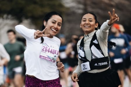 Two women smiling and making peace signs while running in a race as part of community teams, surrounded by other runners. Both wear race bibs and athletic clothing. The background is slightly blurred. Austin Marathon Half Marathon & 5K