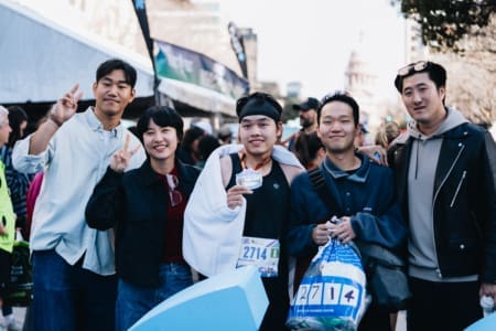 A group of five people, including a runner wearing a medal and race bib, smile and pose together at an outdoor fundraising event with tents and a crowd in the background. Austin Marathon Half Marathon & 5K