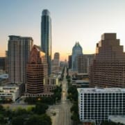 Aerial view of a cityscape at sunrise, featuring tall modern skyscrapers and office buildings lining the Austin Marathon course along a central street with minimal traffic. The sky is clear with a soft, warm glow on the horizon. Austin Marathon Half Marathon & 5K