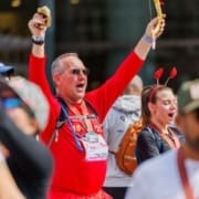 A man in a red outfit with sunglasses cheers with his arms raised at an outdoor destination race in Austin. A woman beside him, wearing red antenna headbands, also appears to cheer. The crowd is festive and energetic. Austin Marathon Half Marathon & 5K