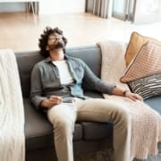 A man with curly hair and glasses reclines on a gray sofa with knitted blankets and patterned pillows, looking relaxed as he enjoys some well-deserved physical recovery, his head tilted back and a remote control in his hand. Austin Marathon Half Marathon & 5K