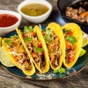 Four hard shell tacos filled with ground beef, diced tomatoes, shredded cheese, and cilantro are arranged on a plate with lime wedges—perfect fuel after that destination race in Austin. Two bowls of salsa, one red and one green, are in the background. Austin Marathon Half Marathon & 5K