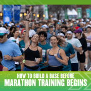 Crowd of runners at a race, two women in front smiling and making peace signs, surrounded by other participants. Text reads: "How to Build a Base Before Marathon Training Begins." Green graphic border emphasizes base building for running success. Austin Marathon Half Marathon & 5K