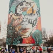 A tall building in Austin features a colorful mural with a woman's face, bold "VOTE" text, and artistic elements. Runners blurred in motion pass by at street level, evoking the spirit of a destination race as leafless trees frame the scene. Austin Marathon Half Marathon & 5K