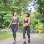 Two young women in athletic clothing are walking and smiling together on a tree-lined path in a park, enjoying a sunny day as they focus on both physical and mental recovery after their recent marathon. Austin Marathon Half Marathon & 5K