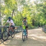 Two people wearing helmets ride bicycles on a sunlit path surrounded by trees. One smiles in front, while the other rides slightly behind along metal railings—an inviting scene found on top running trails and Austin running routes. Austin Marathon Half Marathon & 5K