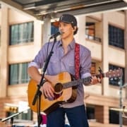 A young man wearing a cap, patterned shirt, and jeans plays an acoustic guitar and sings into a microphone on an outdoor stage in Austin, with tall buildings in the background. Austin Marathon Half Marathon & 5K
