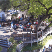 People sit at wooden picnic tables on a large outdoor deck shaded by trees, enjoying Spring in Austin. Some are eating or talking while others, including runners passing by, enjoy one of the city's top patio spots with lively vibes and lush plants. Austin Marathon Half Marathon & 5K