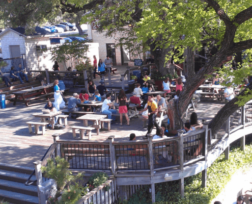 People sit at wooden picnic tables on a large outdoor deck shaded by trees, enjoying Spring in Austin. Some are eating or talking while others, including runners passing by, enjoy one of the city's top patio spots with lively vibes and lush plants. Austin Marathon Half Marathon & 5K