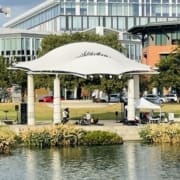 A white outdoor pavilion with musicians performing underneath it, surrounded by plants and water in the foreground—an inviting spot along Austin running routes, with modern office buildings and trees framing the spring running scene. Austin Marathon Half Marathon & 5K