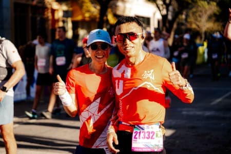 Two smiling runners from the Friends and Family Teams, wearing red shirts, sunglasses, and race bibs, pose with thumbs up at an outdoor race event, surrounded by other participants and trees in the background. Austin Marathon Half Marathon & 5K
