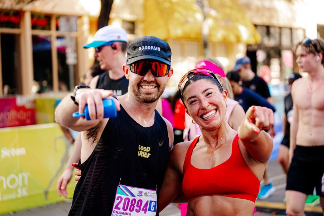 Two smiling runners, a man in a black tank top and cap and a woman in a red sports bra, point at the camera after finishing a race—perfect for Friends and Family Teams or team building group activities, with fellow participants and a colorful outdoor backdrop. Austin Marathon Half Marathon & 5K