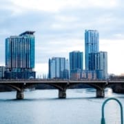 A city skyline with tall modern buildings rises behind a bridge spanning a wide river under a cloudy sky. The crowded bridge and riverbank trees capture the vibrant energy that makes Austin a top destination race location for many reasons. Austin Marathon Half Marathon & 5K