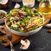 A fresh salad in a white bowl with greens, avocado, olives, broccoli, seeds, and creamy dressing—perfect for physical recovery—surrounded by mushrooms, seeds, and a bottle of olive oil on a dark table. Austin Marathon Half Marathon & 5K
