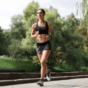 A woman in athletic wear jogs on a paved path outdoors, surrounded by green trees and grass on a sunny day, focusing on marathon training and building her running preparation. Austin Marathon Half Marathon & 5K