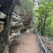 A paved trail runs alongside a rocky cliff, bordered by a metal railing, with dense green trees and foliage on the right side—one of the top running trails perfect for spring running among Austin running routes. Austin Marathon Half Marathon & 5K
