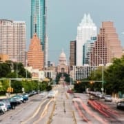 A view of downtown Austin, Texas, featuring cars with blurred lights on a city street, modern skyscrapers, and the Texas State Capitol building in the background—part of the iconic Austin Marathon course under a cloudy sky. Austin Marathon Half Marathon & 5K