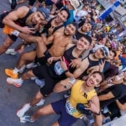A group of runners poses for a photo at a crowded street race, celebrating post marathon joy. They smile, make peace signs, and one shirtless man stands in the middle. Most wear athletic gear and race bibs, with other runners behind them. Austin Marathon Half Marathon & 5K