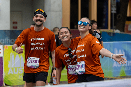 Three smiling runners in matching orange shirts reading "I THOUGHT THIS WAS A BAR CRAWL?" pose happily at a race event, looking like school teams or collegiate teams with their arms around each other and race bibs proudly displayed. Austin Marathon Half Marathon & 5K