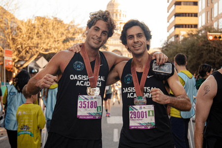 Two runners in “Oasis Futsal Academy” tank tops, proudly wearing medals and race bibs, smile after a race. Behind them, fellow participants and a domed building set the scene—echoing the camaraderie found in collegiate teams. Austin Marathon Half Marathon & 5K
