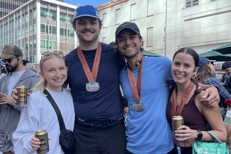 Four smiling people stand close together outdoors, proudly wearing running medals around their necks and holding cans, likely after completing a race with their social teams. Buildings and other participants are visible in the background, celebrating their training efforts. Austin Marathon Half Marathon & 5K