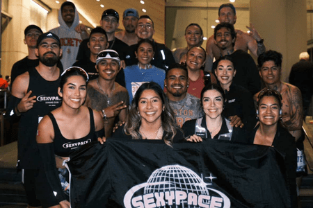 A group of smiling runners from social teams pose together indoors, holding a black "SEXY PACE" flag with a globe graphic. Most wear athletic clothing and race bibs, showing off their training spirit with peace signs and playful gestures. Austin Marathon Half Marathon & 5K