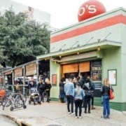 People stand in line outside a green and red restaurant with a large red ball on the roof displaying "O'S." Bicycles are parked in front, and trees provide shade nearby in one of the vibrant neighborhoods along the Austin Marathon course. Austin Marathon Half Marathon & 5K