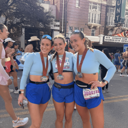 Three women wearing matching light blue crop tops and blue shorts stand together smiling with race bibs and medals, celebrating their achievements in the Austin Marathon team program at an outdoor street event with other participants in the background. Austin Marathon Half Marathon & 5K